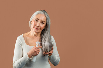 Smiling mature caucasian female with natural beauty applies spray on gray hair, looking at camera