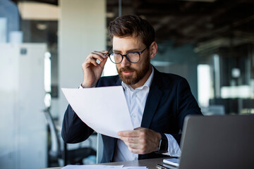 Focused middle aged businessman working with papers in modern office interior, reading financial documents