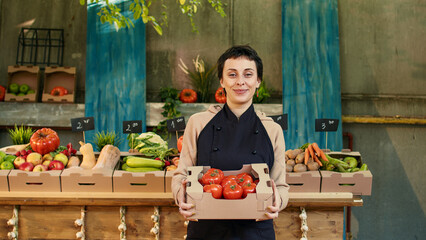 Woman farmer holding box of fresh bio tomatoes at healthy food marketplace, selling homegrown...