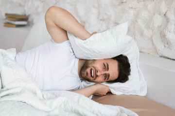 Tired angry sad young caucasian guy lies on bed, covers his ears with pillow, suffers from noisy neighbors