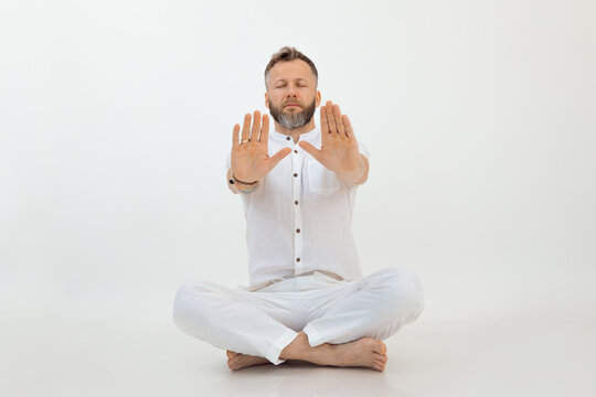 Calm middle-aged bearded man sitting with crossed legs on floor, stretching hands with open palms on white background.