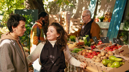 Female stall holder greeting loyal client and hugging at food marketplace, being cheerful selling locally grown produce. Positive farmer sharing hug with customer, enjoying visit at street festival.