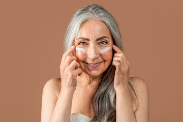 Glad elderly caucasian female with natural beauty applies cream on her face, fight with wrinkles