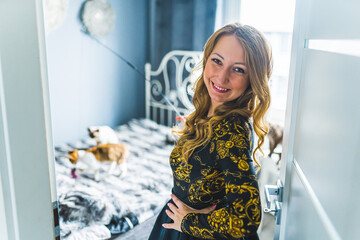 Shoulder level shot of a blonde woman with her hands on her hips looking at the camre and smiling. Devon rex playing on the bed in the background. Blurred background. High quality photo