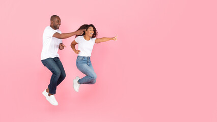 Fototapeta na wymiar Excited black man and woman jumping up in the air and pointing at free space, pink studio background, panorama