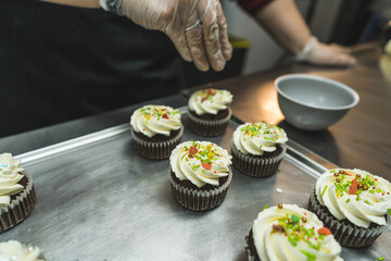 A baker wearing a transparent gloves decorating cupcakes on a baking tray. Cupcakes with white...