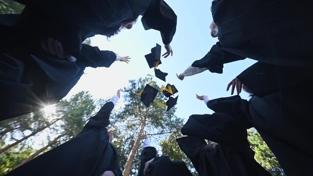 Classmates in graduation gowns toss their hats outdoors. Bottom view. 