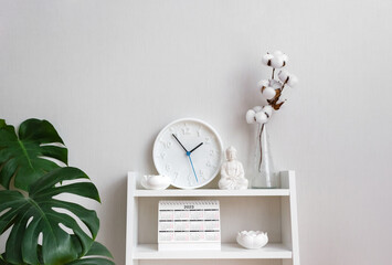 Buddha figurine, clock, lotus candlesticks and table calendar surrounded by monstera and vases with branch of cotton on white shelf. Concept mental health and recreation. Selective focus, copy space