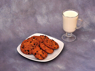 A glass of hot milk and a plate of several oatmeal chocolate chip cookies on a gray abstract background.