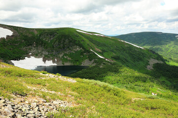 Fototapeta na wymiar A small lake in a high-altitude hollow with the remains of snow on the slopes overgrown with grass.