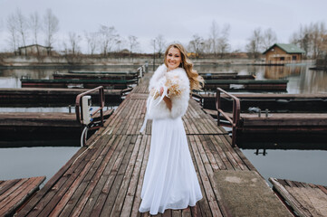 Fototapeta premium A beautiful smiling curly blonde bride in a white dress, a fur coat walks along a wooden pier in autumn, against the background of the river. Wedding photography, portrait.