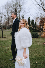 A stylish bearded groom in a black coat and a beautiful smiling curly blonde bride in a white dress, fur coat with a bouquet of reeds are walking outdoors in the park. Wedding photography, portrait.