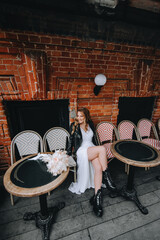 Beautiful curly smiling blonde bride, biker, lover of musical rock in a black leather jacket, white dress sits on chairs in the city on the street. wedding photography.