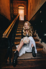 A cool bride, biker, rock lover in a black leather jacket, white dress with a bouquet sits on the steps indoors against the background of a wall with a chandelier. wedding photography.