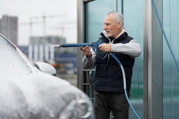 Caucasian bearded grey-haired elderly man, a driver using a pressurized water hose sprays a jet of...