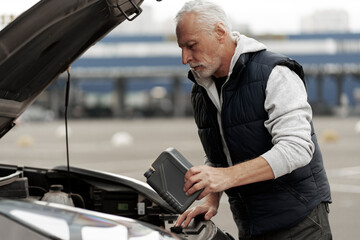 Caucasian bearded senior man driver, standing by his car with open hood  checking the motor oil level and adding the lubricant oil in the engine. Auto service and car repair concept 