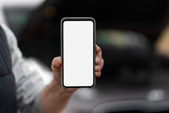 Close up of male hand holding modern smartphone with blank screen copy advertising space for mobile app, against blurred background of broken down car with open hood. Car service concept