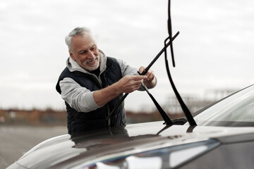 Handsome smiling senior Caucasian man, driver installing new windshield wipers by himself at outdoors parking lot. Automobile industry. Safety driving. Car maintenance. People. Auto service concept  - obrazy, fototapety, plakaty