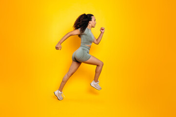 Fototapeta na wymiar Full length side view photo of a fitness sporty brazilian or hispanic woman in sportswear, training working out, jumping, running, looking aside, isolated orange background. Sports lifestyle