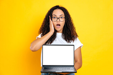 Shocked amazed brazilian or latino young curly woman, wearing basic white t-shirt, holding open laptop with blank white mock-up screen, looks excited at camera, standing on isolated yellow background