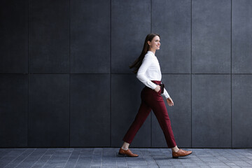 Fototapeta na wymiar Young woman in formal clothes walking near grey wall outdoors, space for text