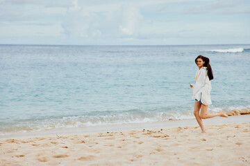Sports woman runs along the beach in summer clothes on the sand in a yellow T-shirt and denim shorts white shirt flying hair ocean view