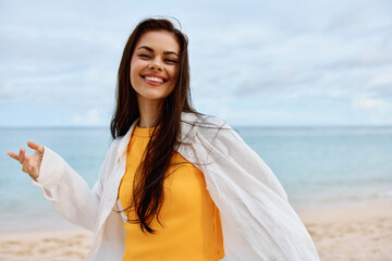 Happy woman smile with teeth with long hair brunette walks along the beach in a yellow t-shirt denim shorts and a white shirt near the sea summer journey and feeling of freedom, balance