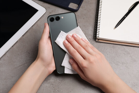 Woman wiping smartphone with paper towel at gray table, closeup
