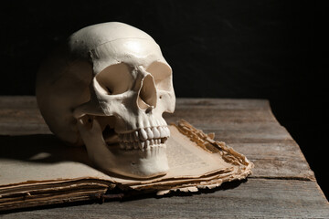Human skull and old book on wooden table against black background, space for text