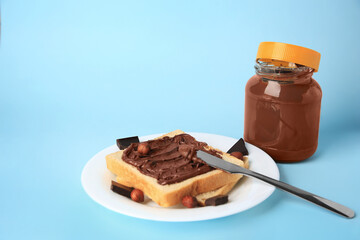 Tasty toast with chocolate paste and nuts near jar of cream on light blue background