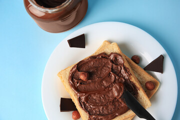 Spreading chocolate paste onto bread near jar of cream on light blue background, flat lay