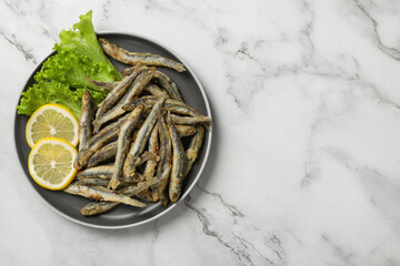 Plate with delicious fried anchovies, lemon and lettuce leaves on white marble table, top view. Space for text