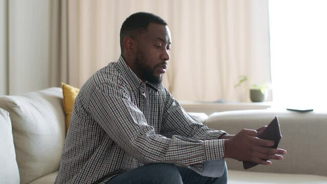 Financial crisis. Unemployed african american man sitting at home with empty wallet, feeling lack of money, empty space