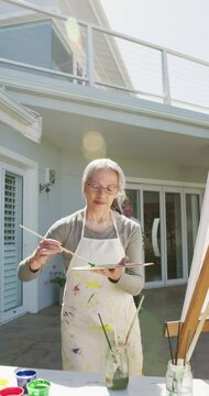 Vertical video of happy asian senior woman painting in garden on sunny day