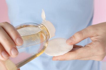 Woman pouring micellar water from bottle on cotton pad against pink background, closeup