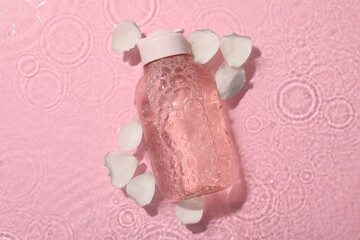 Wet bottle of micellar water and petals on pink background, top view