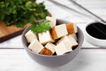 Bowl of smoked tofu cubes, soy sauce and parsley on white wooden table, closeup
