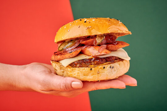 mano de una mujer sosteniendo una hamburguesa con chorizo carne y pan con ajonjoli en un fondo de varios colores verdes y rojo
