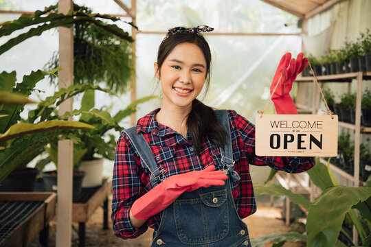 Portrait of beautiful smiling Asian woman looking at camera, holding open wooden sign board " Welcome we are OPEN in the greenhouse. Shop and Store for sell plants