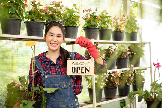 Portrait of beautiful smiling Asian woman looking at camera, holding open wooden sign board " Welcome we are OPEN in the greenhouse. Shop and Store for sell plants