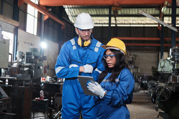 Two industrial workers in safety uniforms and hardhats, male manager, and Black colleague work with tablet to check metalwork machines in manufacturing factory. Professional production engineer team.