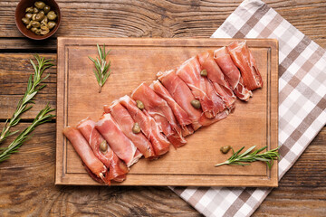 Cutting board with slices of tasty ham, rosemary and capers on wooden background