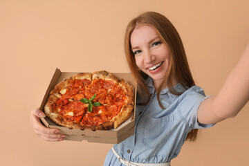 Young woman with box of tasty pizza taking selfie on beige background