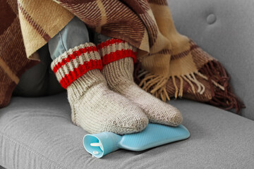 Young man warming his feet with hot water bottle on sofa at home, closeup
