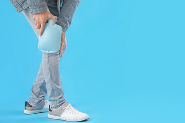 Young man warming his knee with hot water bottle on blue background