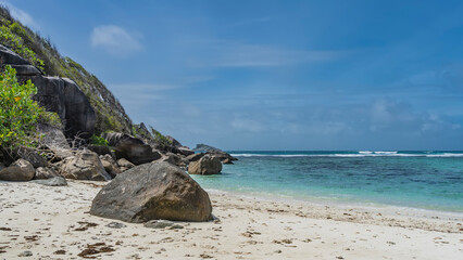 Obraz premium Secluded tropical beach. Boulders on the sand and at the water's edge. Foam of waves in the turquoise ocean. The slope of a green hill against a blue sky. Seychelles. Moyenne Island.