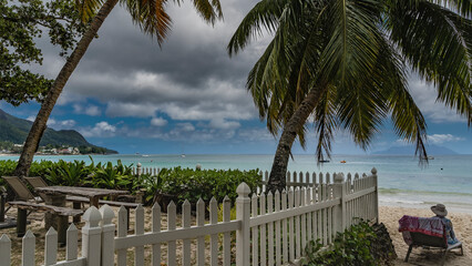 In the recreation area on a tropical beach, behind a white fence, there is a wooden table, benches....