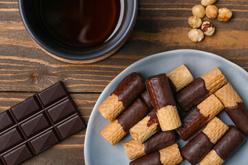 Plate with tasty wafer rolls, chocolate and cup of coffee on wooden background, closeup