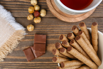 Plate with tasty wafer rolls, chocolate, cup of tea and nuts on wooden background, closeup