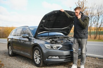 Man repairing a broken car by the road. Man having trouble with his broken car on the highway roadside. Man looking under the car hood. Car breaks down on the autobahn. Roadside assistance concept.
